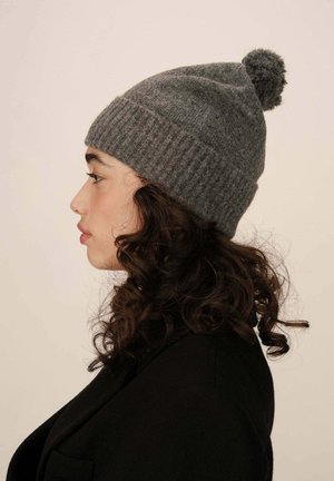 Femme de profil avec des cheveux bouclés portant un bonnet en tricot gris foncé avec pompon et un manteau noir devant un fond neutre.