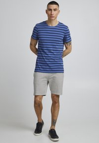 T-shirt à rayures bleues et noires à manches courtes, associé à un short gris. Le modèle porte des sneakers noires et affiche des tatouages sur les bras et les jambes.