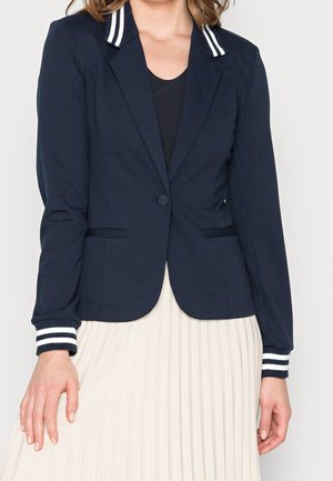Femme portant un blazer bleu marine avec col et poignets à rayures blanches par-dessus un haut noir, assorti à une jupe plissée blanche.