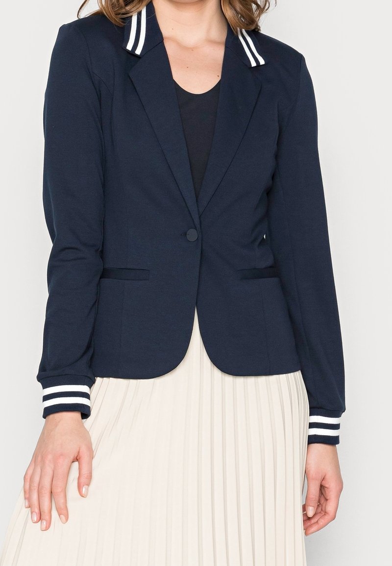 Femme portant un blazer bleu marine avec col et poignets à rayures blanches par-dessus un haut noir, assorti à une jupe plissée blanche.