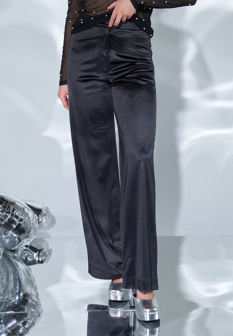 Personne portant un pantalon large noir brillant, un top noir transparent avec des détails en perles et des talons compensés argentés métalliques, debout sur une surface réfléchissante.