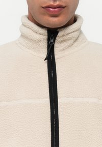 Beige fleecejakke med høy krage, med svart glidelås og kontrasterende sømmer. Myk tekstur og minimalt design.