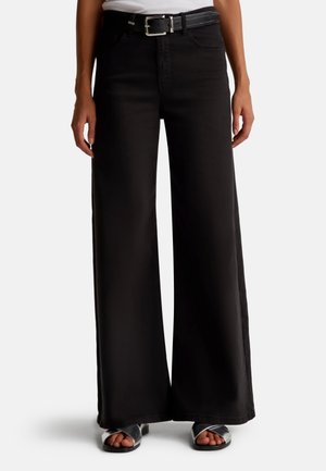 Person iført sort bukser med høj talje og vide ben, med sort bælte og sølvfarvede sandaler, stående mod en hvid baggrund.