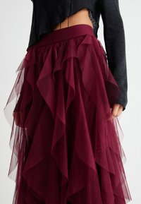 Burgundy tulleskirt som har lager av volanger, elastisk midjeband och en genomskinlig textur. Synlig svart stickad topp som överlappar vid midjan.
