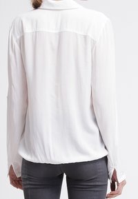 Blusa bianca a maniche lunghe in un tessuto leggero e morbido; dettaglio sulla spalla dietro; polsini sagomati; orlo curvo; design minimalista.
