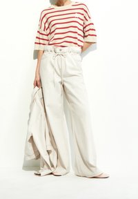 Osoba w wysokich, szerokich kremowych spodniach z wysokim stanem oraz krótkim czerwono-białym swetrze, trzymająca pasującą kremową kurtkę.