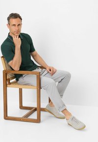 Dunkelgrünes Poloshirt mit weißen Akzenten, hellgraue Hose und hellgraue Schuhe. Modell sitzt auf einem Holzstuhl mit geflochtenem Sitz.
