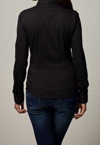 Maglietta nera a maniche lunghe con un design attillato, texture liscia e polsini con bottoni, abbinata a jeans blu scuro con dettagli di cucitura sottili.