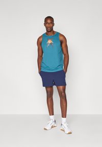 Teal tank topp med grafisk logo, marineblå shorts, og hvite joggesko med lyse brune såler. Modellen står mot en ensfarget bakgrunn.