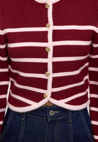 Cárdigan a rayas rojas y blancas con escote redondo, cinco botones dorados con emblema de león y diseño corto. Combinado con pantalones vaqueros oscuros.