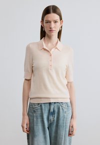 Ανοιχτό ροδακινί κοντομάνικο πλεκτό πόλο με γιακά, που διαθέτει ημίσειο ρεμπέτικο και διακριτή ριμπ ύφανση, σε συνδυασμό με ανοιχτό τζιν παντελόνι.