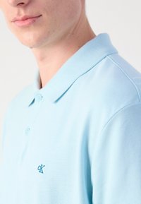Lyseblå polo-shirt lavet af struktureret stof. Har en klassisk krave og tre knapper, samt et lille broderet logo på brystet.