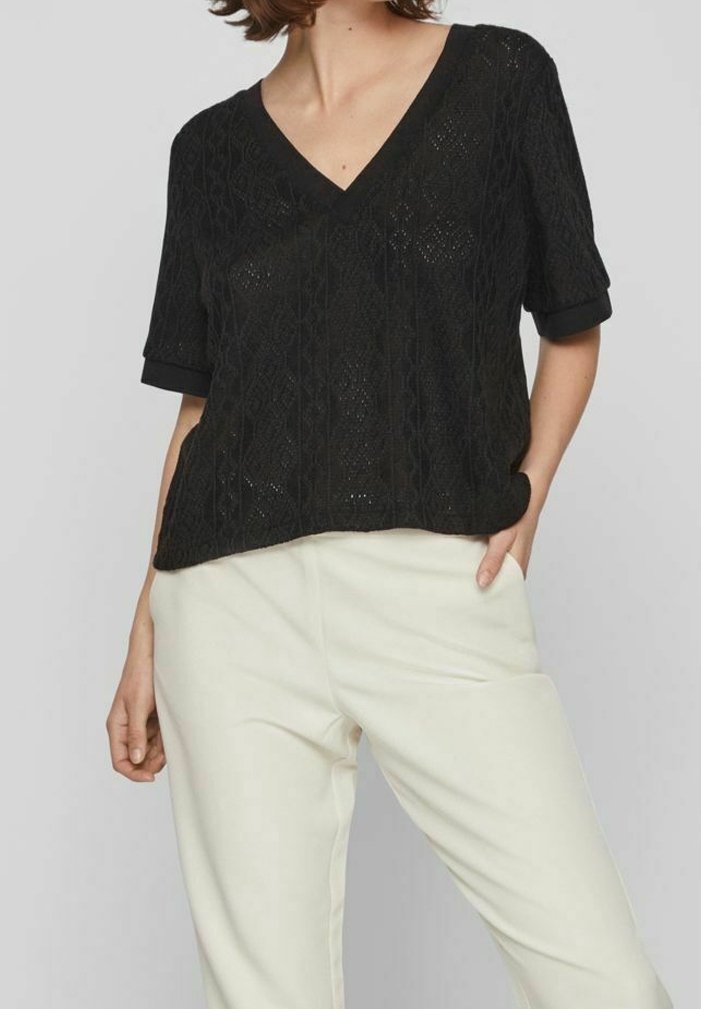 Femme portant un haut texturé noir à manches courtes et col en V, ainsi qu'un pantalon blanc taille haute, une main dans la poche et l'autre détendue.