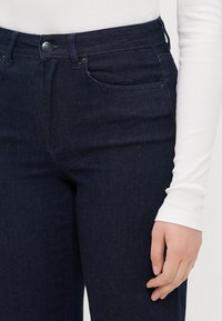 Tumma farkut sisältävät korkeaa vyötäröä, hienovaraisia ompeleita ja klassisen viisi-taskun muotoilun. Pintakuvio näyttää sileältä vähäisellä kulutuksella.