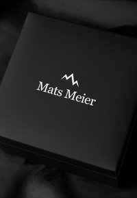 Boîte noire avec une finition mate lisse, présentant le texte blanc "Mats Meier" et un simple logo de montagne. Design minimaliste sans accents supplémentaires.