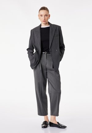 Femme debout portant un blazer oversize gris foncé et un pantalon large assorti, chemise noire, ceinture noire et mocassins noirs sur fond blanc.