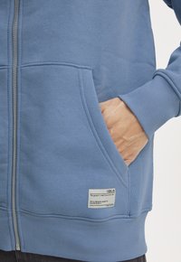 Sudadera de cremallera azul claro, hecha de un tejido suave, con un bolsillo tipo canguro y puños acanalados. Etiqueta adjunta al dobladillo con instrucciones de cuidado.