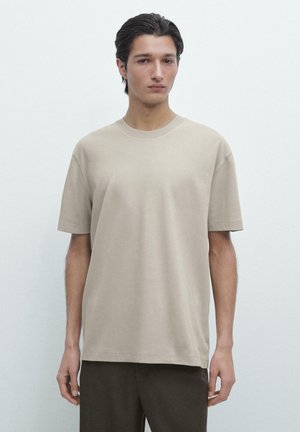 Young man with dark hair wears beige short-sleeve T-shirt and dark pants, standing against plain light gray background.