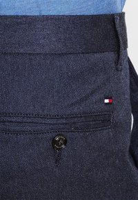 Närbild av marinblå byxor med bakficka, svart knapp och en liten röd, vit och blå Tommy Hilfiger-logotyp nära fickan.