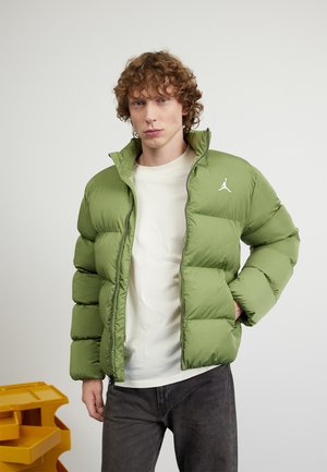 Lichtgroene pufferjack met een hoge kraag, ritssluiting en wit logo op de linkerkant van de borst. Draag het over een wit shirt en grijze broek.