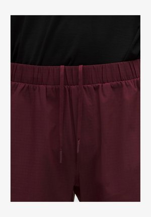 Közelkép a bordó rugalmas derékpántos nadrágról, elöl zsinórral, fekete felsővel viselve.