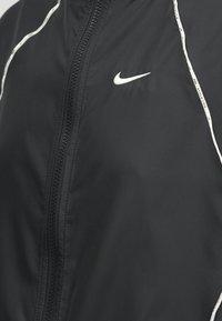 Μαύρο μπουφάν Nike με φερμουάρ, λευκές ρίγες στους ώμους και λευκό λογότυπο Nike Swoosh στο αριστερό στήθος.