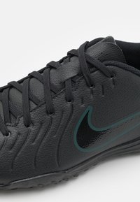 Zapatilla deportiva negra con cuero sintético texturizado, diseño de cordones y un sutil logo de Nike en negro y teal. Patrón de suela circular.