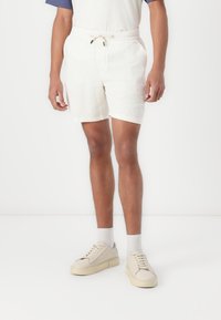 Pantalones cortos blancos texturizados con una cintura elástica y cordón. Combinados con zapatillas y calcetines en tonos neutros, sobre un fondo liso.