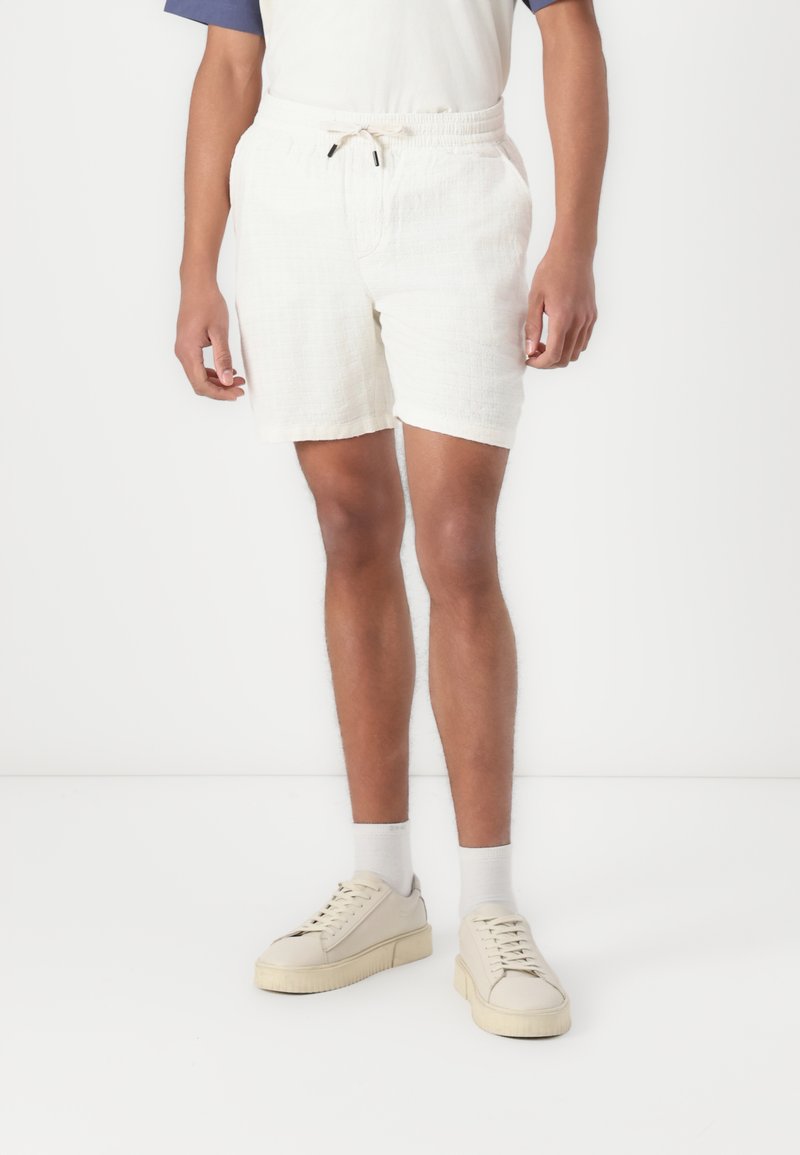 Pantalones cortos blancos texturizados con una cintura elástica y cordón. Combinados con zapatillas y calcetines en tonos neutros, sobre un fondo liso.