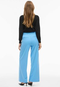 Lyseblå bukser med vide ben og elastisk talje, med sidelommer, båret med sorte hælede sko og en sort top.