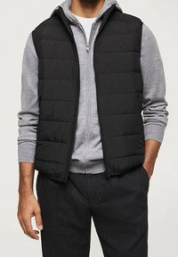 Schwarze Steppweste mit gestepptem Design, getragen über einem grauen, zip-up Pullover. Die Weste ist ärmellos, hat einen Stehkragen und einen Reißverschluss.