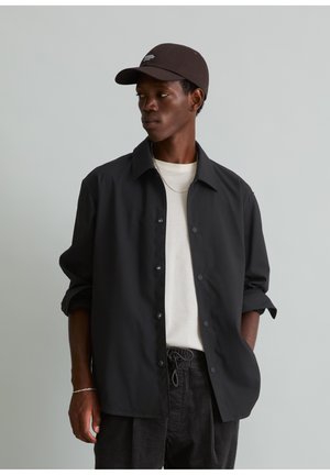 Jonge man die een zwart shirt draagt over een wit T-shirt, zwarte broek, een zwarte pet en een zilveren ketting, kijkt opzij met zijn handen in zijn zakken.