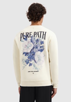 Man met krullend haar, gekleed in een crèmekleurige sweatshirt met blauwe bloemenpatronen en de tekst "PURE PATH" en "OWN THE JOURNEY" op de achterkant, zwarte broek.