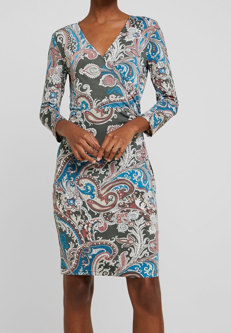 Femme portant une robe longue jusqu’aux genoux, à manches longues, imprimé cachemire bleu, marron et blanc, avec un décolleté en V, mains jointes de manière détendue à la taille.