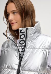 Srebrna kamizelka z wysokim kołnierzem i czarnym zamkiem błyskawicznym z logo "JOOP!". Materiał jest błyszczący i pikowany w poziome sekcje.