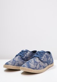 Chaussures décontractées bleues à lacets avec un motif de feuilles blanches et des semelles tissées beiges, exposées sur une surface blanche.