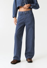 Personne portant un pantalon large à rayures verticales bleues et blanches, une veste courte assortie et des baskets marron, debout devant un fond blanc.
