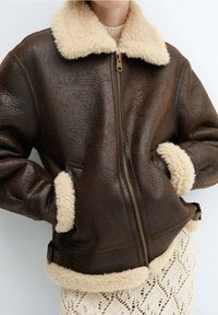 Femme portant une veste en cuir marron foncé avec un col et des manches en shearling crème, les mains dans les poches, sur une jupe en tricot crème avec un motif en dentelle.