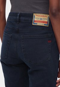 Personne portant un jean denim bleu foncé de la marque Diesel avec des poches arrière et un patch en cuir marqué sur la ceinture.