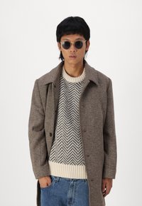 Brūns houndstooth mētelis ar ieliektu apkakli, kombinēts ar rakstainu krēmkrāsas un pelēku džemperi un zilu džinsu biksēm. Apaļas saulesbrilles.
