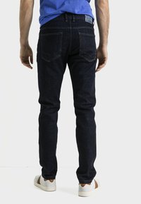 Jeans de mezclilla azul oscuro con un corte ajustado, que cuentan con bolsillos traseros con detalles de costura, combinados con zapatillas de color claro.