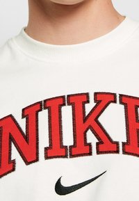 Vit sweatshirt med djärv röd "NIKE"-text med svarta detaljer. Mjuk tyg med ribbad rundhals och synliga sömdetaljer.