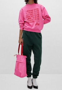 Felpa rosa con la scritta "LOVERS CLUB", abbinata a pantaloni della tuta verdi. Una borsa rosa con manici rossi. Le scarpe col tacco nere completano l'outfit.