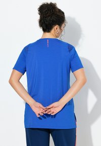 Žena s kudrnatými vlasy svázanými nahoře má na sobě modré sportovní tričko s krátkými rukávy a tmavě modré kalhoty, stojí s rukama sepnutýma za zády.