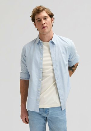 Młody mężczyzna w jasnoniebieskiej koszuli z podwiniętymi rękawami, noszący kremowy t-shirt i jasnoniebieskie dżinsy, stojący na tle jednolitego jasnoszarego tła.