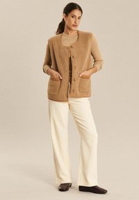 Braune Fleece-Weste mit rundem Ausschnitt, vorderen Knöpfen und zwei aufgesetzten Taschen, getragen über einem beigen Oberteil und crèmefarbenen weiten Hosen.