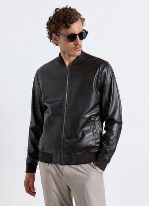 Mann trägt schwarze Lederjacke, beige Hose und schwarze Sonnenbrille, schaut zur Seite mit einer Hand in der Tasche.