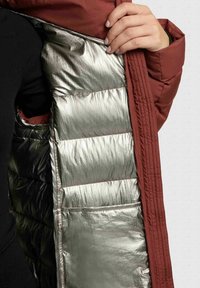 Intérieur d'une doudoune bordeaux avec une doublure argentée brillante, une texture matelassée et un rembourrage gonflé avec des détails noirs.