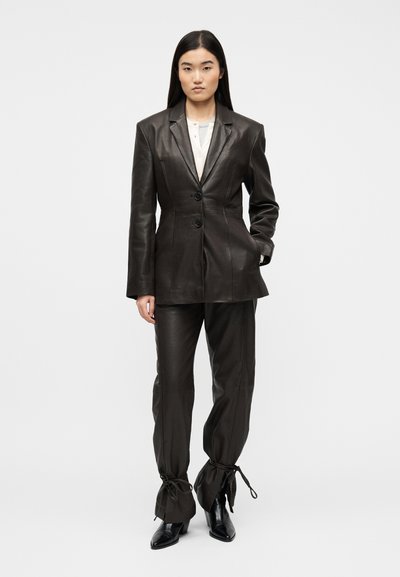 Femme portant un blazer en cuir noir et un pantalon assorti noué aux chevilles, associé à des bottes à talons noires, debout devant un fond blanc.