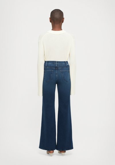 Persona in piedi di spalle, che indossa un maglione a coste color crema e jeans a vita alta larghi di un blu scuro con tacchi.